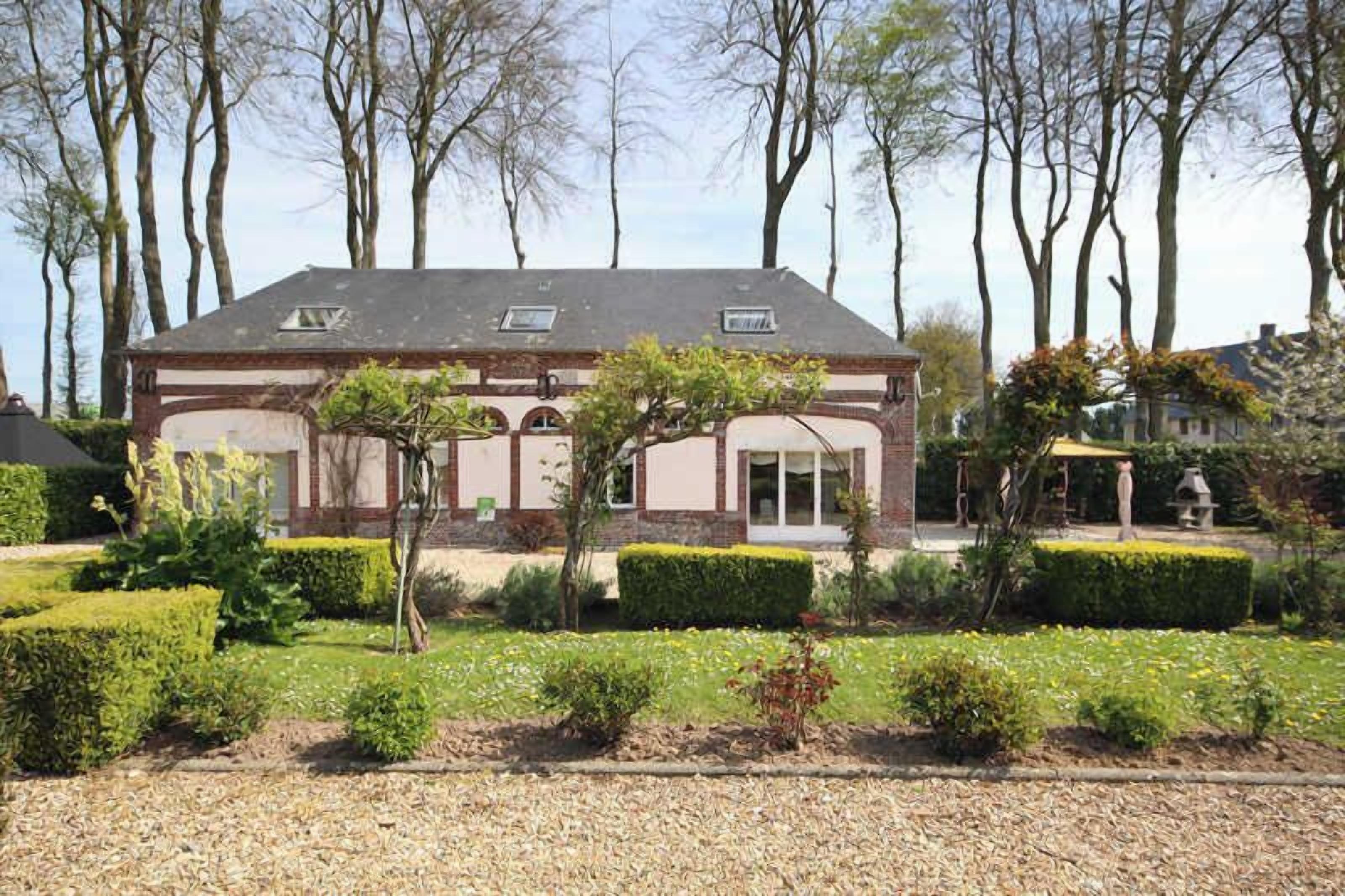 Une maison à deux étages, dotée d’une façade symétrique, d’une entrée centrale et d’un jardin bien entretenu agrémenté de haies et d’arbres.