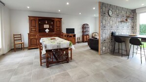 Un grand espace de salle à manger avec une table et des chaises en bois, un buffet intégré en bois, et un coin cuisine doté d’un comptoir-bar et de tabourets.