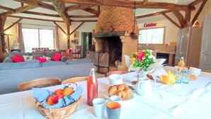 A rustic room with exposed wooden beams, a brick fireplace, a dining table set with breakfast, and a cozy seating area.