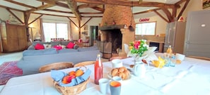 Une pièce rustique avec des poutres apparentes, une cheminée en briques, une table à manger dressée pour le petit-déjeuner et un coin salon confortable.