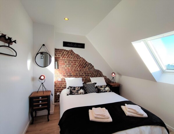 Ein Schlafzimmer mit schräg laufender Decke, einer Ziegelwand als Blickfang, einem Bett mit schwarz-weiß gemustertem Überwurf, zwei Nachttischlampen, einem kleinen Holfnachtschränkchen und einem Spiegel an der Wand.