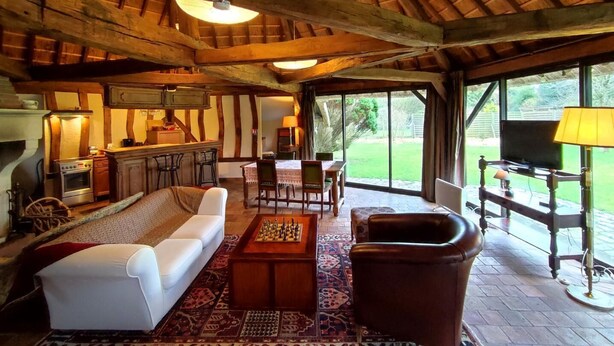 Un interior rústico con vigas de madera a la vista, chimenea, comedor y un amplio ventanal con vistas a un jardín.