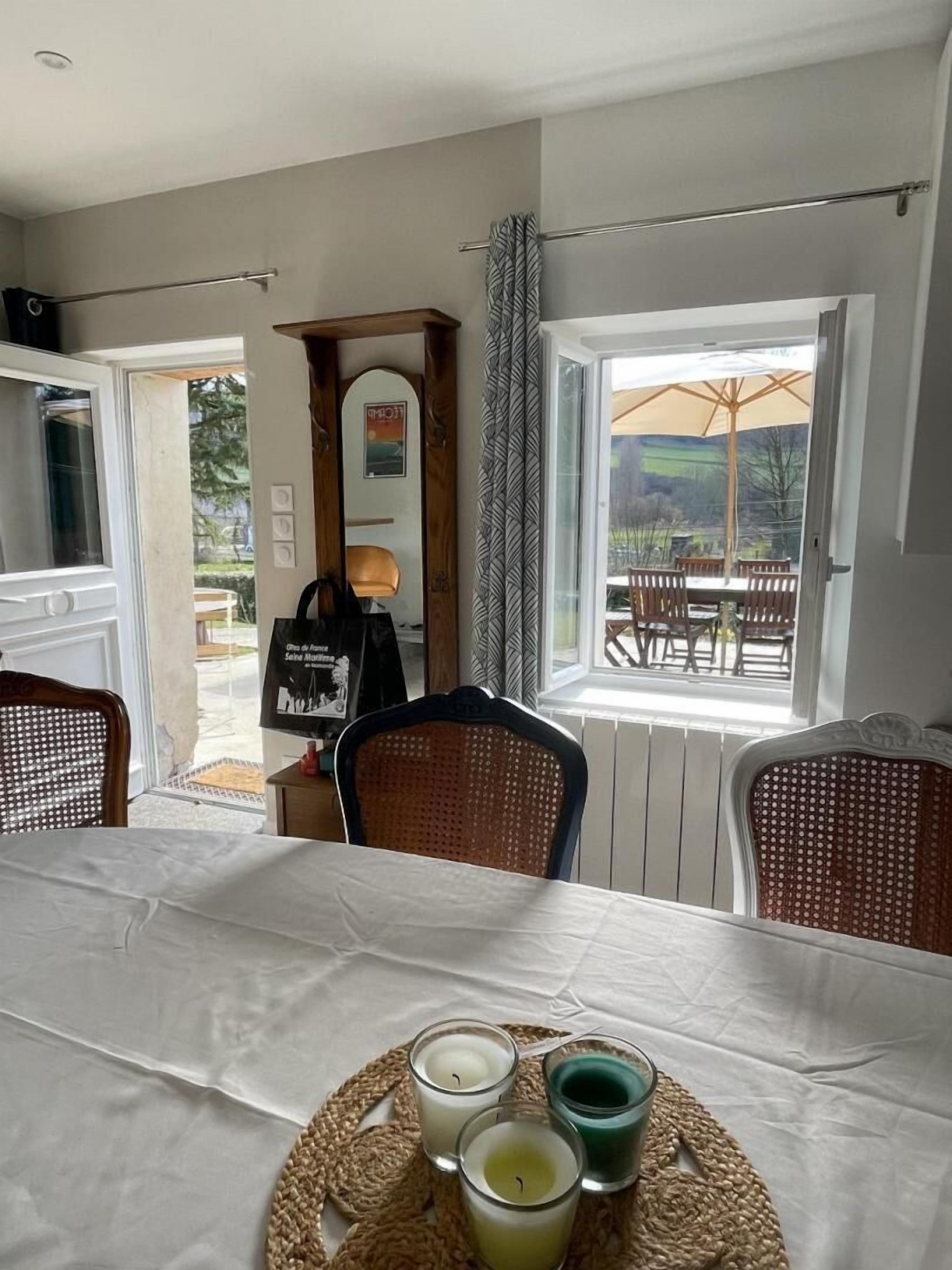 Uma área de jantar com mesa posta para quatro pessoas, vista do exterior por uma grande janela e bandeja de vime com velas.