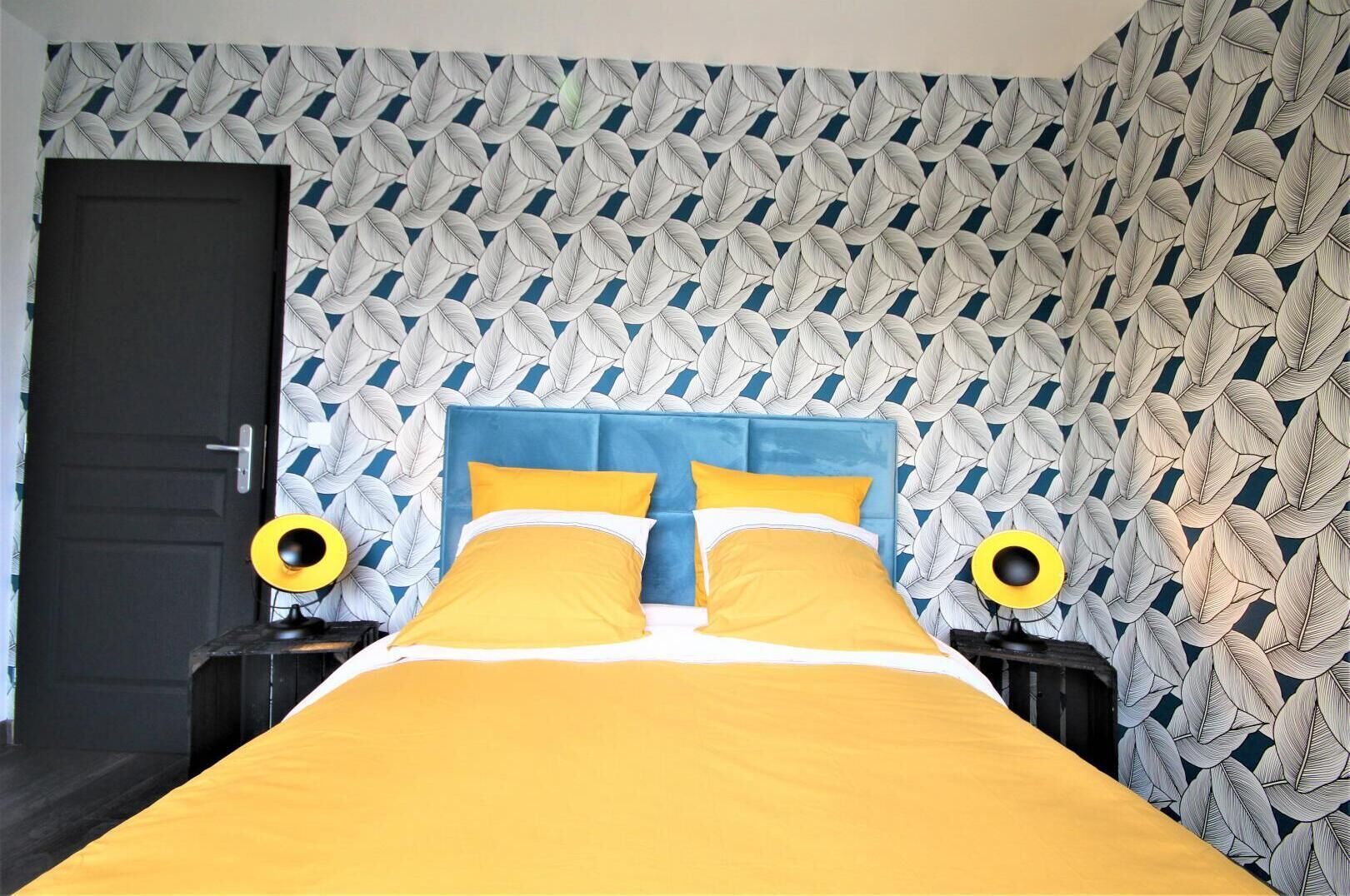 Una cama con cabecera azul y ropa de cama amarilla, flanqueada por dos mesitas de noche con lámparas amarillas. La pared presenta un diseño geométrico de hojas.