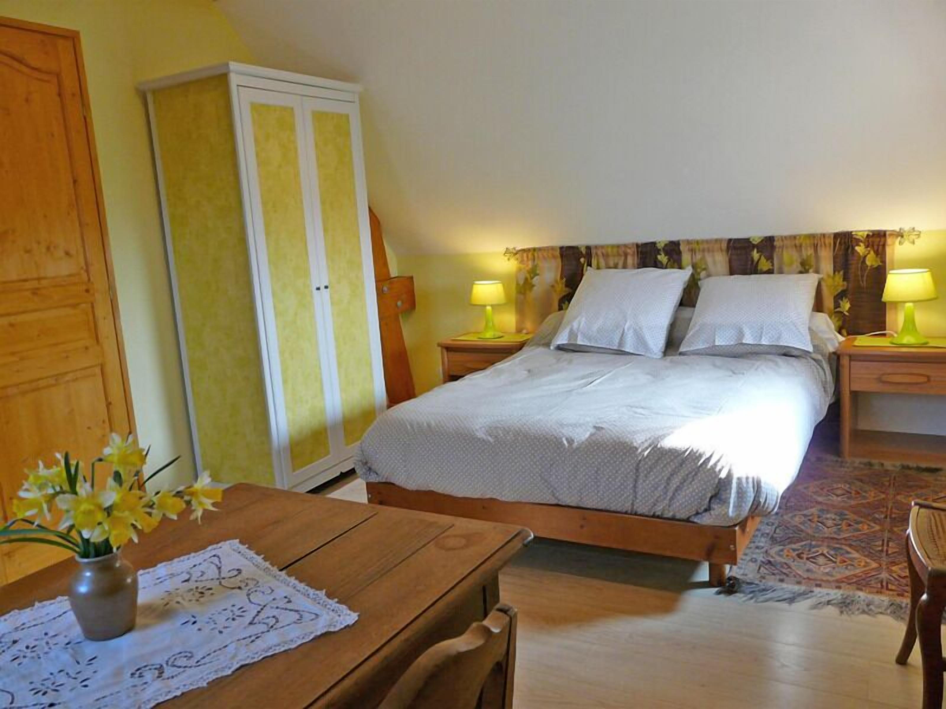 A bedroom with a bed, two bedside tables, a wooden wardrobe, a table with a vase of flowers, and a patterned rug.