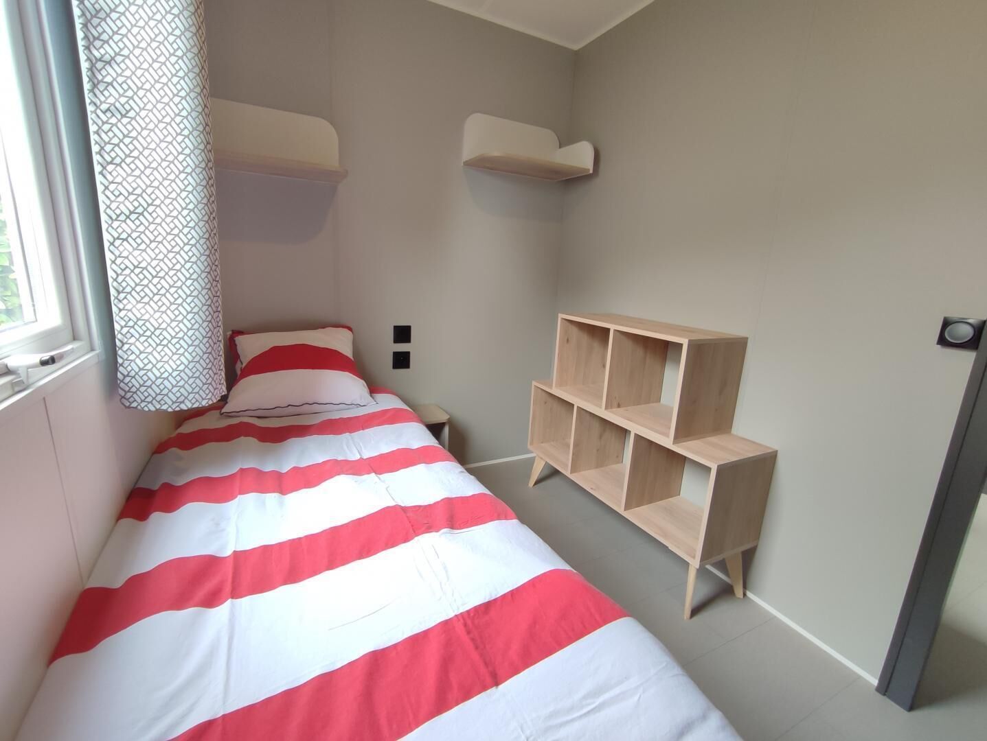 Quarto pequeno, com uma cama de solteiro, colcha listrada, estante de madeira e janela com cortinas estampadas.