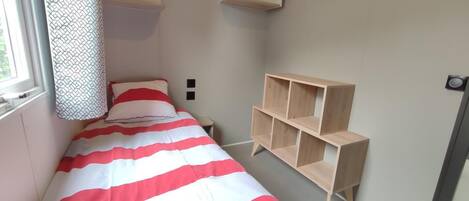 Quarto pequeno, com uma cama de solteiro, colcha listrada, estante de madeira e janela com cortinas estampadas.