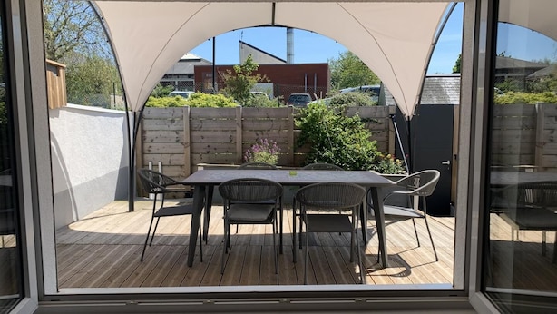 En overdækket terrasse med bord og stole, træterrasse og udsigt til en murstensbygning og grøntområde.