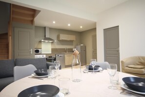 Un coin repas avec une table ronde dressée pour quatre personnes, une cuisine en arrière-plan et un canapé gris.