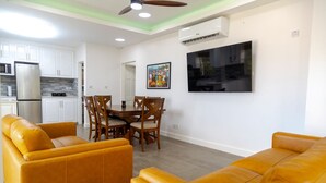 Ein modernes Wohnzimmer mit gelben Sofas, einem Esstisch mit Stühlen, einer Küchenzeile mit weißen Schränken und einem wandmontierten Fernseher.