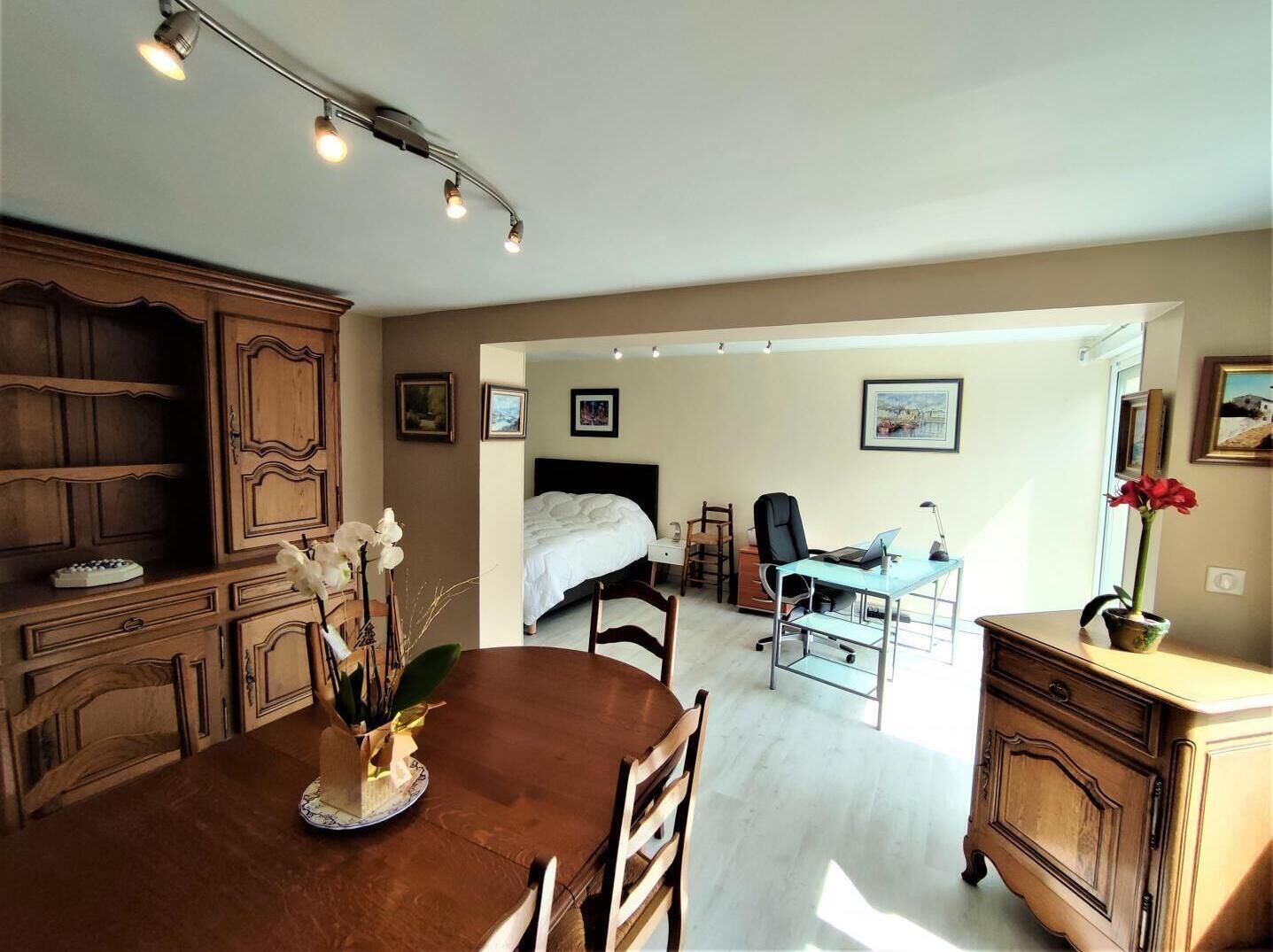 A hotel room with a wooden dining table, a bed with a white duvet, a desk with a laptop, and a chair. There are framed pictures on the wall and a vase with flowers on the dresser.