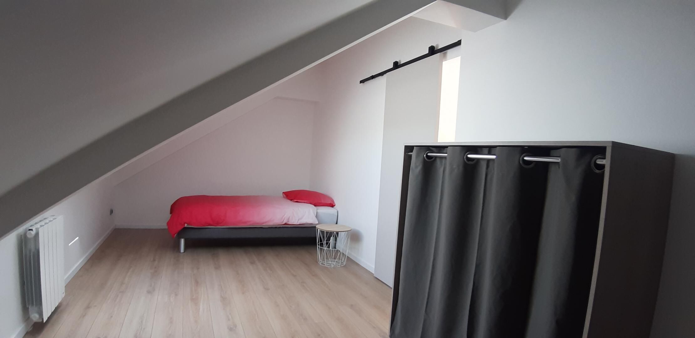 Una habitación pequeña con techo inclinado, una cama, una mesita redonda y una cortina negra.