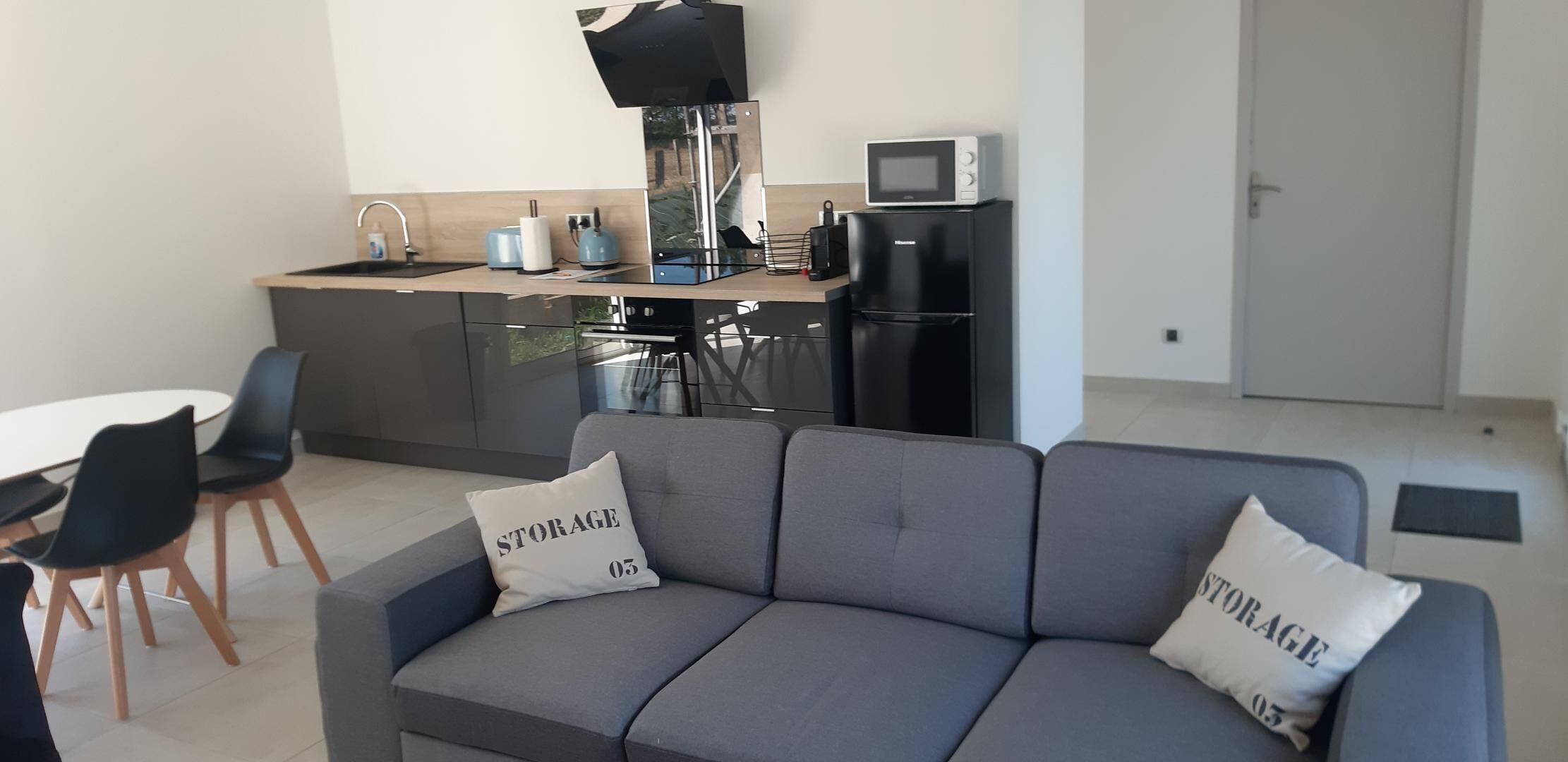 Un salon moderne avec un canapé gris, deux coussins décoratifs, une petite table à manger avec des chaises et un coin cuisine équipé d’un micro-ondes et d’une bouilloire.