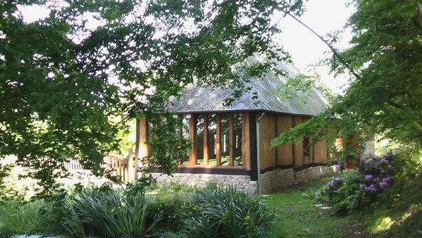 Un gazebo con base in pietra e pilastri in legno, immerso nel verde e con un piccolo stagno.