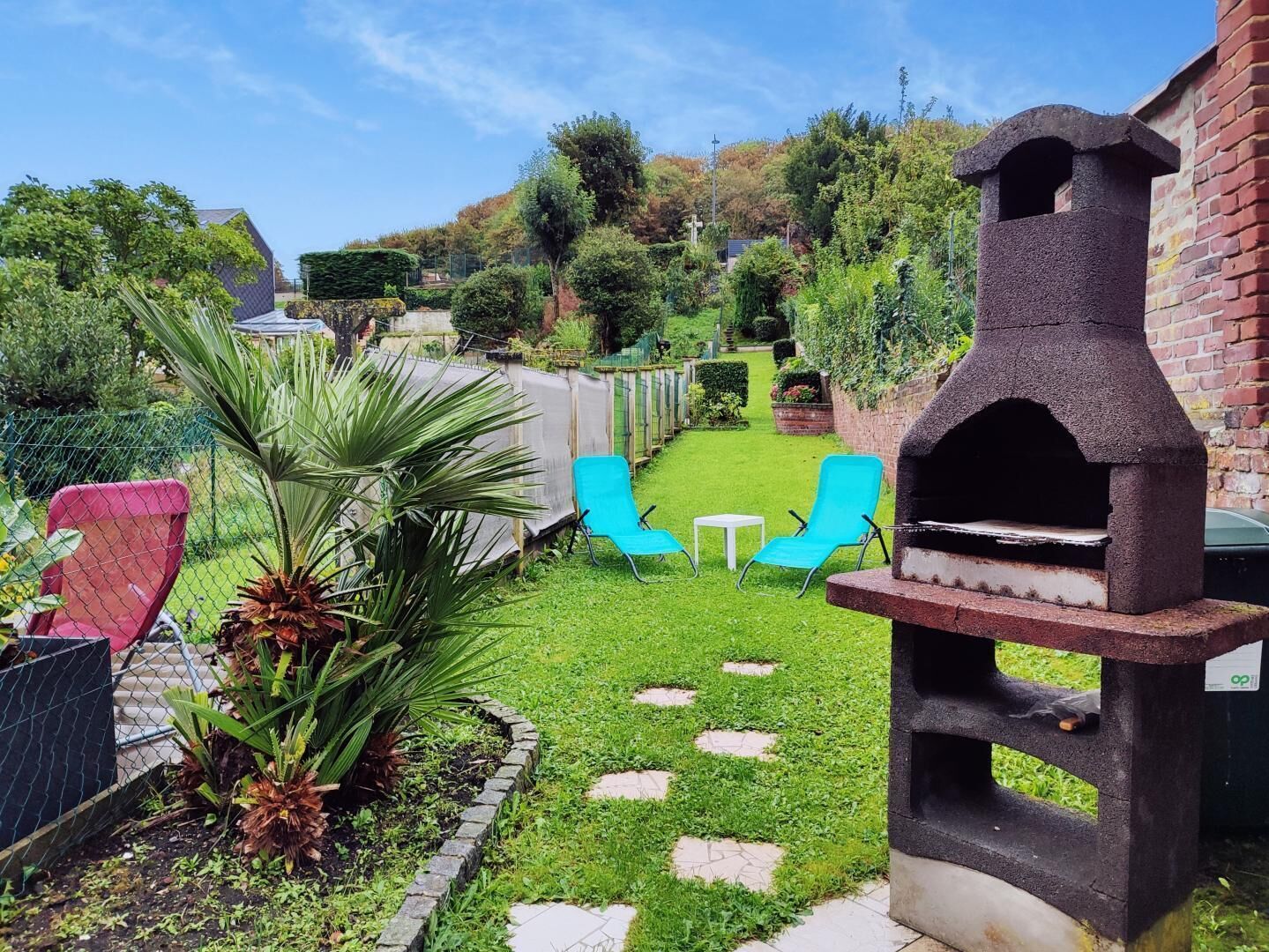 A backyard with a barbecue grill, two blue chairs, a small table, and a garden with a palm plant.