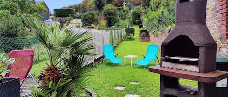 A backyard with a barbecue grill, two blue chairs, a small table, and a garden with a palm plant.