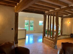 A spacious room with exposed wooden beams, a dining area with green chairs, and a view of the outdoors through a glass door.