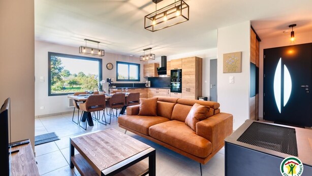 A modern living room with a sofa, coffee table, and dining area with chairs. A kitchen with wooden cabinets and a window is visible in the background. - Gîtes de France® - In the heart of Senneville (SENNEVILLE SUR FECAMP)