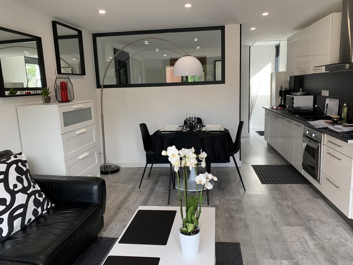 A modern kitchen with white cabinets, a dining area with a black table and chairs, and a living area with a black sofa and a coffee table.