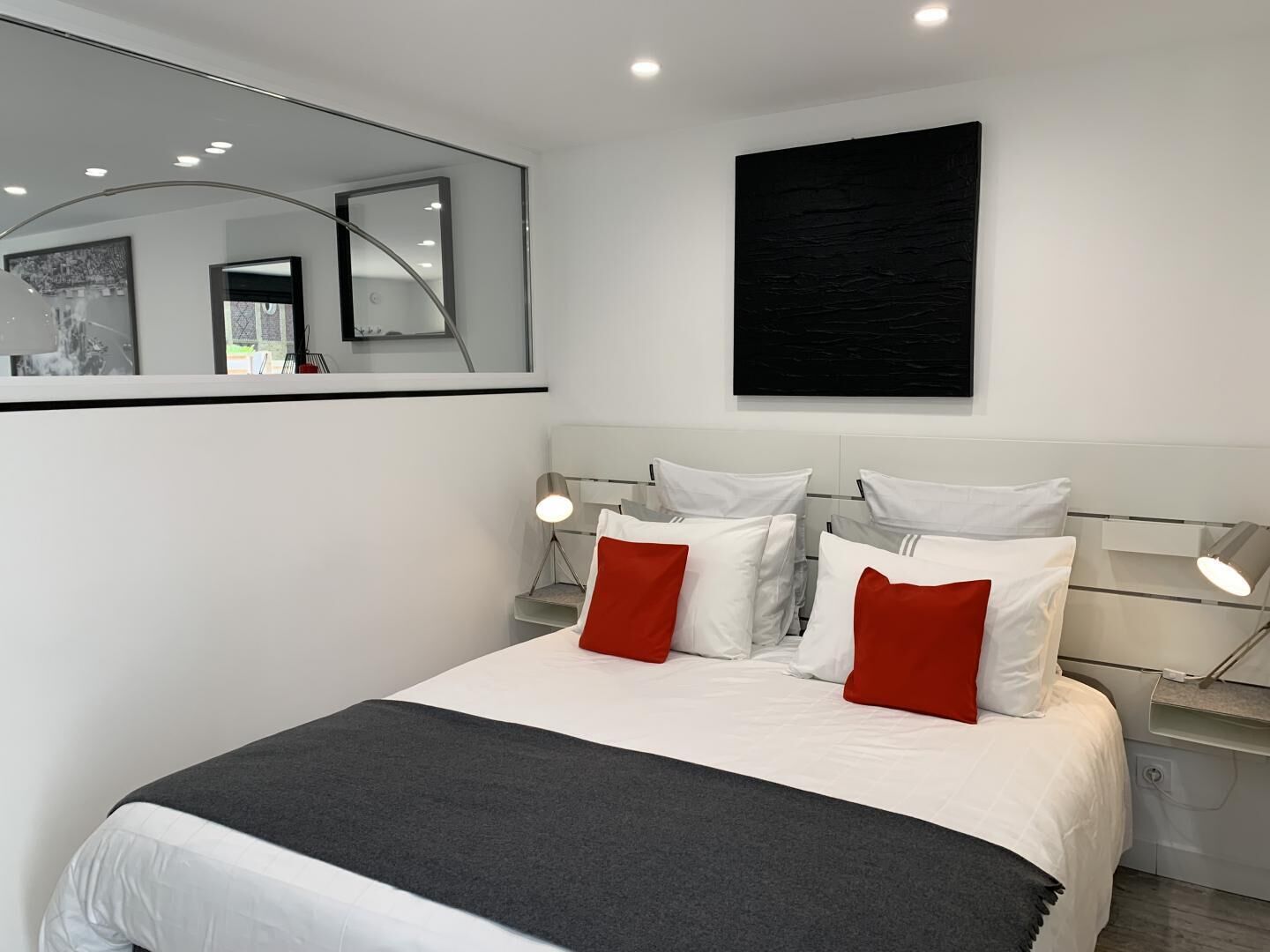 Une chambre moderne avec un grand lit, deux tables de chevet avec des lampes, un grand miroir et une œuvre d’art noire accrochée au mur.