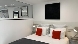 Une chambre moderne avec un grand lit, deux tables de chevet avec des lampes, un grand miroir et une œuvre d’art noire accrochée au mur.