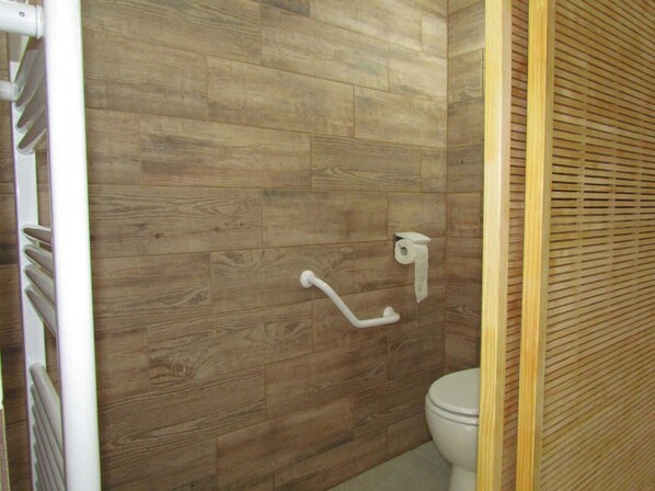 A bathroom with wooden flooring, a white toilet, a handrail, and a towel dispenser.