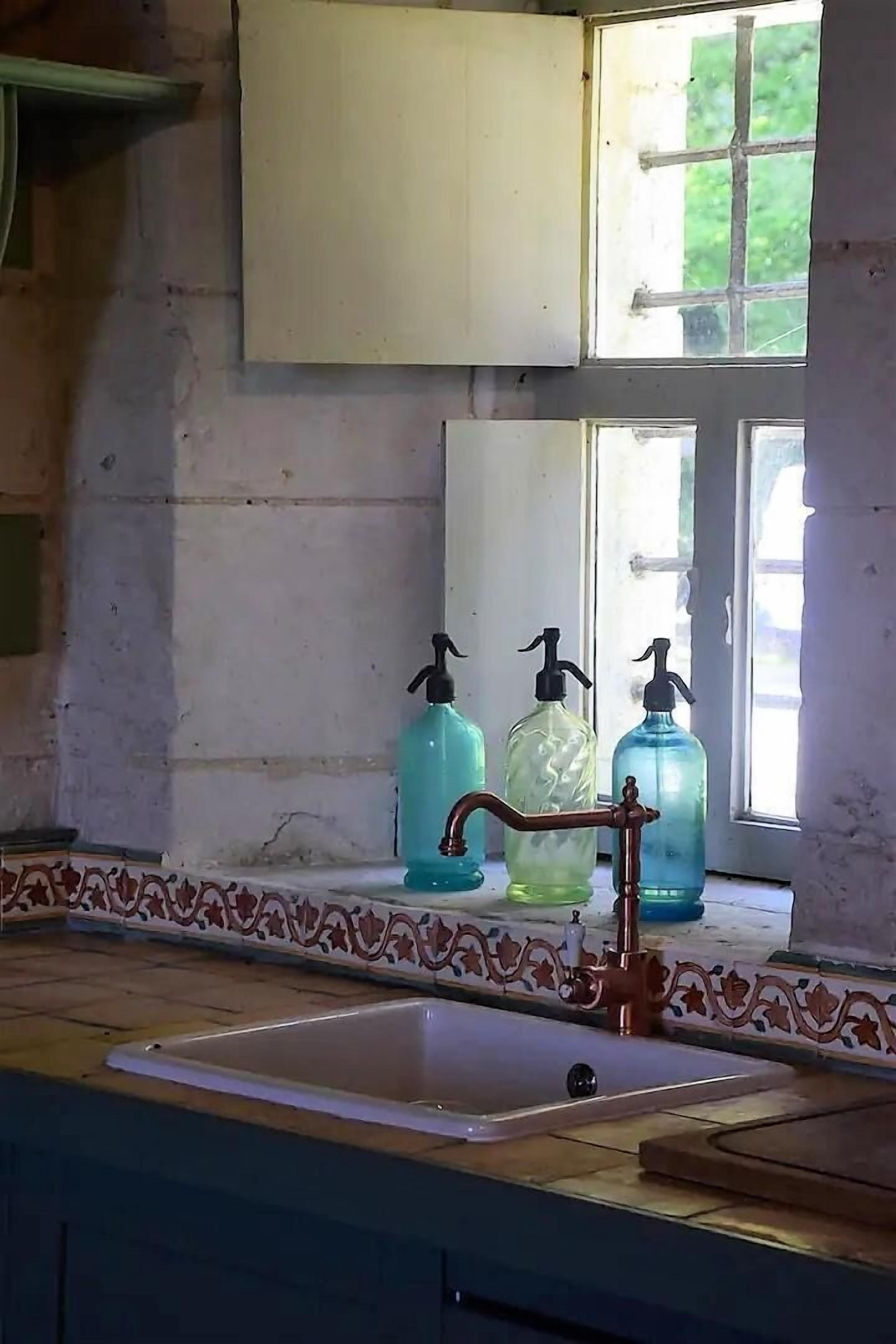 A kitchen with a sink, three hand soap dispensers, and a window with a view of greenery.