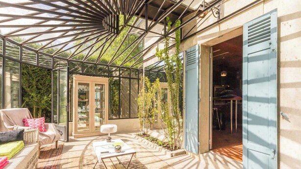 A conservatory with a glass ceiling, a seating area with a sofa and armchairs, a coffee table, and a door leading to an interior space.