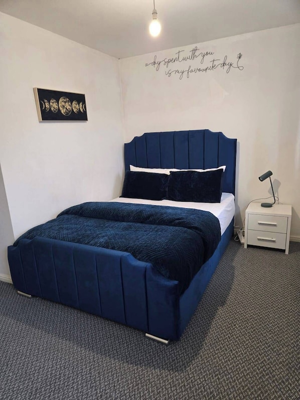 A bedroom with a dark blue bed, white bedding, a small bedside table with a lamp, and a wall with a moon phase artwork. - West Winds Sleeps 4 With Garden (Wolverhampton)