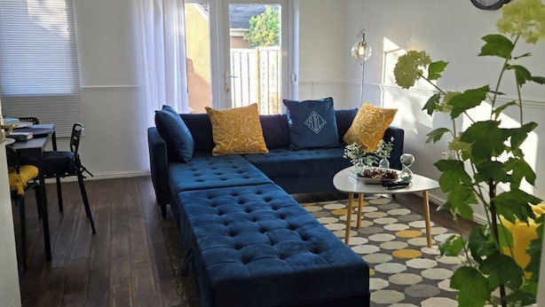 Un soggiorno con un divano blu scuro, un tavolino rotondo e un tappeto decorato. C'è anche una zona pranzo con tavolo e sedie, e una finestra con persiane.