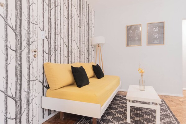 Un dormitorio moderno con una cama blanca, ropa de cama amarilla, una alfombra estampada y una pequeña mesa blanca con un florero.