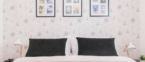 Una cama bien hecha con edredón blanco y negro, dos almohadas negras y una blanca. Encima de la cama, hay tres carteles enmarcados en la pared.