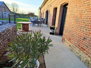 A patio area with a round table and chairs, potted plants, and a brick building with windows. - Gîtes de France® - L'Hirondelle (TURRETOT)