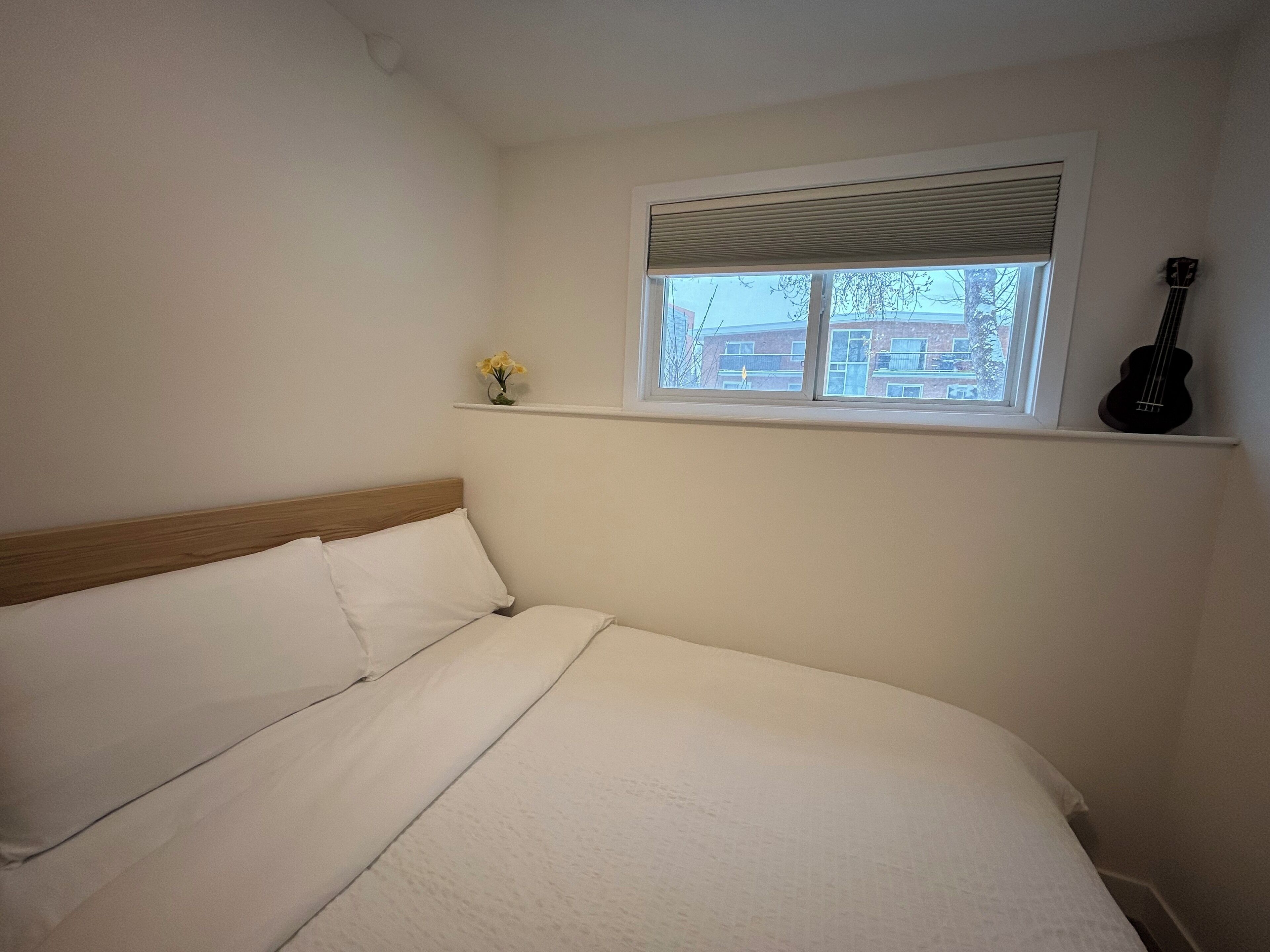 Une chambre à coucher avec un lit, une fenêtre avec des stores, une petite étagère sur laquelle est posé un vase, et une guitare appuyée contre le mur.
