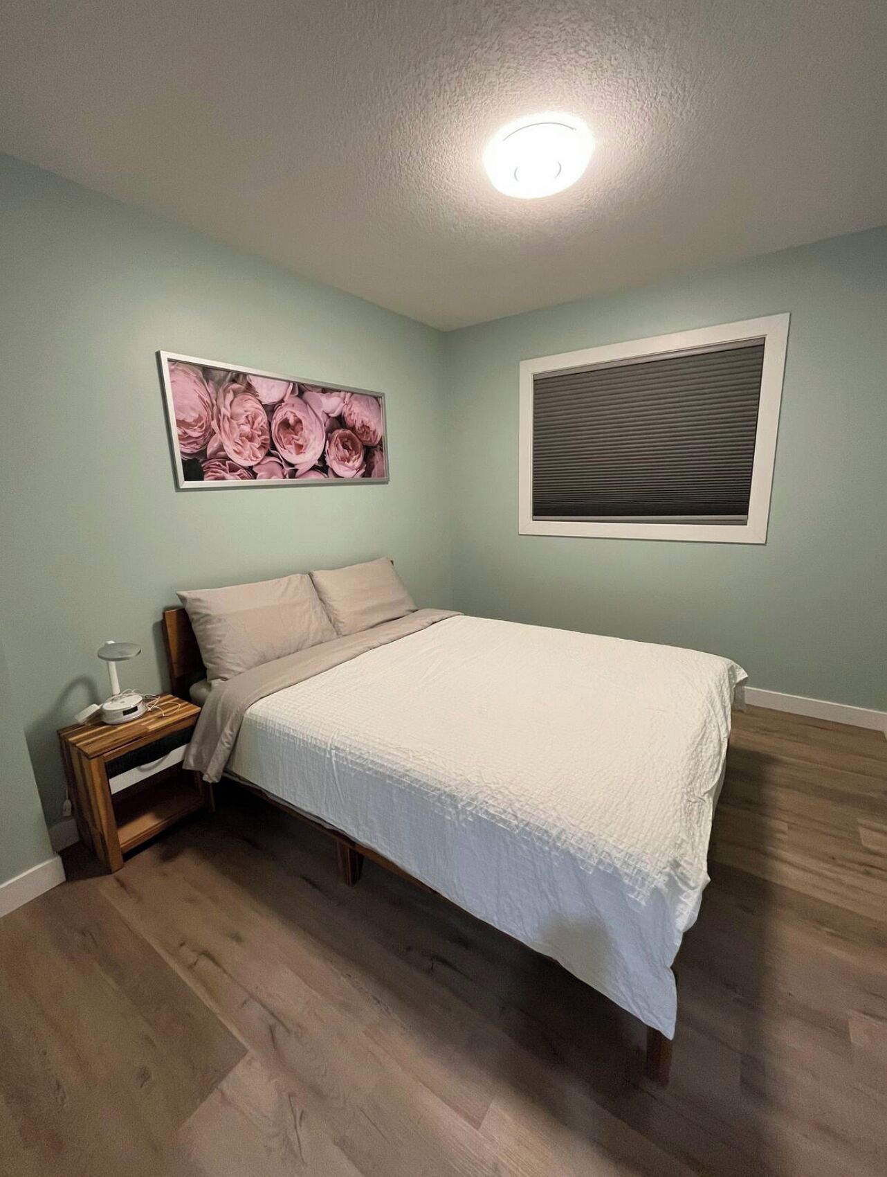 Une chambre à coucher comprenant un lit, une table de chevet, un tableau représentant des fleurs accroché au mur, une fenêtre avec des stores et un luminaire au plafond.