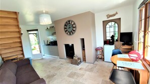 A living room with a sofa, a dining table with chairs, a fireplace, a kitchen in the background, and a large wall clock. - Gîtes de France® - La Normandeuse (REALCAMP)