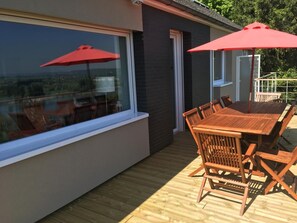 A wooden deck with a table and chairs, a red umbrella, and a large window overlooking a landscape.