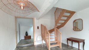 Una habitación amplia con piso de concreto, una escalera de madera, una silla blanca, una mesita pequeña y una lámpara colgante.