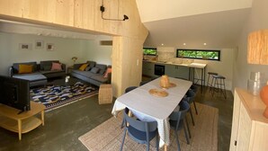Un interior moderno con comedor estilo loft, una cocina con ventanales amplios y un espacio de sala con sofá y televisor.