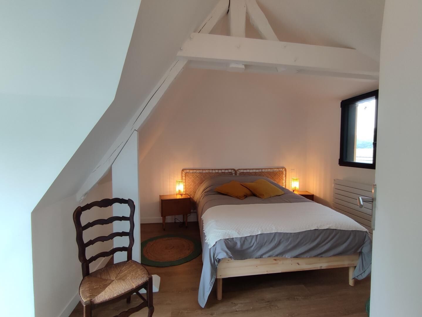Een slaapkamer met een schuine plafond, houten bedframe, een stoel, twee bedlampen en een raam.