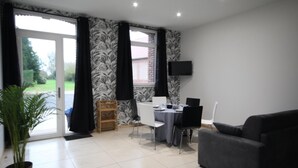 A modern living room with a sofa, dining table, and chairs. There is a large window with curtains, a flat-screen TV mounted on the wall, and a potted plant. - Gîtes de France® - Louis-Philippe (BAROMESNIL)