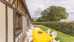 An outdoor dining area with a yellow table and white chairs, a stone-paved patio, and a view of a hedge and trees.