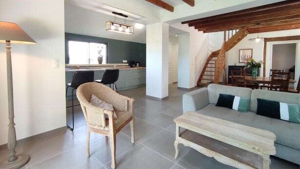 Un vaste espace de vie comprenant un canapé, un banc en bois et un fauteuil en osier. Un escalier mène à une salle à manger avec une table et des chaises.