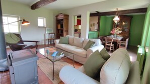 Ein gemütliches Wohnzimmer mit einer Couch, Sesseln, einem Couchtisch und einem Bücherregal. Es gibt einen Essbereich mit Tisch und Stühlen. Das Zimmer verfügt über freiliegende Holzbalken an der Decke und grüne Wände.