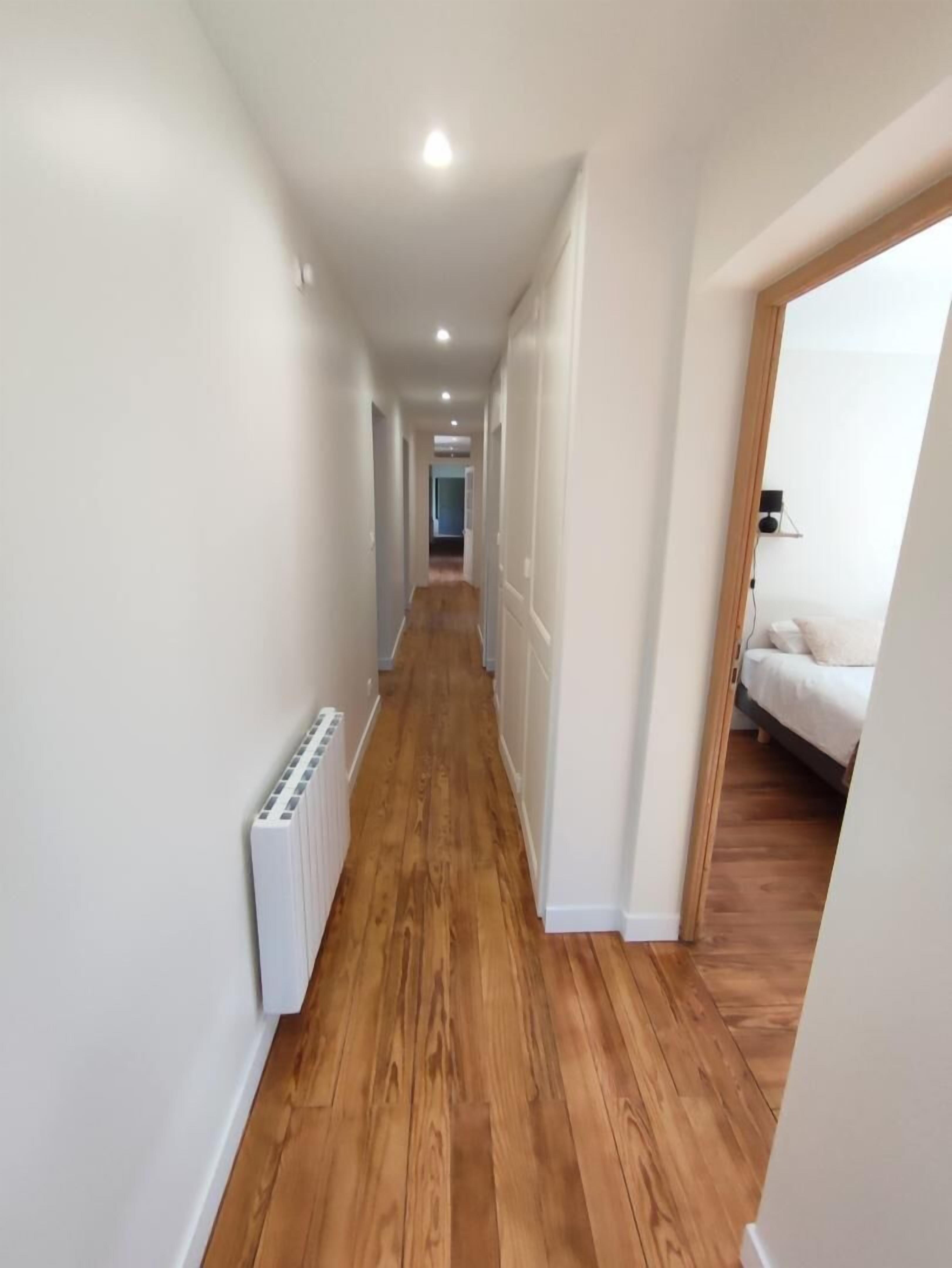 A narrow hallway with wooden flooring and white walls, leading to a bedroom with a bed and a nightstand.