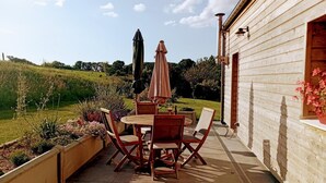 Un patio con mesa y sillas de madera, rodeado de plantas en macetas y vistas a un campo verde.