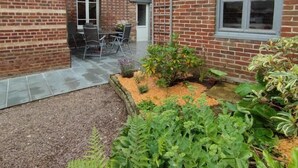 A brick house with a patio area, outdoor seating, and a garden with various plants. - Gîtes de France® - Gîte du Dun (FONTAINE LE DUN)