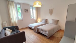 A room with two beds, a sofa, a hanging light fixture, and a window with curtains. - Gîtes de France® - Gîte du Dun (FONTAINE LE DUN)