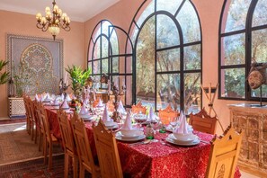 A dining area with a long table set for a meal, large arched windows, and a decorative wall with intricate patterns.