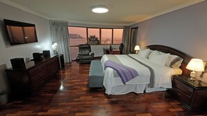 A hotel room with a large bed, a desk, a chair, a TV, and a view of the sea.